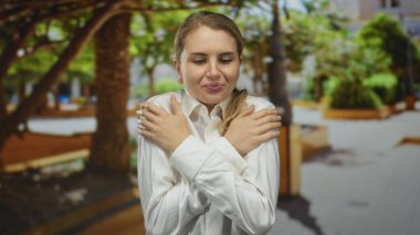 Yeşil bir parkta üşüyen bir kadın beyaz bir gömlek giyiyor, sarı saçlı, açık havada, doğada bir ürperti anı yakalıyor..