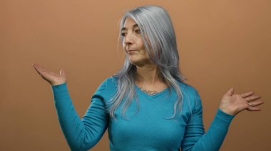 Mavi kazaklı yaşlı kadın, izole edilmiş kahverengi arka plan duvarına el hareketi yaparak rahat ve düşünceli bir davranış sergiliyor..