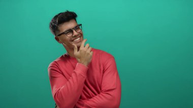 Genç bir adam pembe bir kazakla canlı yeşil bir arka plana karşı güvenle gülümsüyor. Neşeli ve rahat bir atmosfer sergiliyor. İspanyol mirasını vurguluyor..