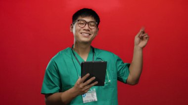 Gülümseyen adam yeşil önlük giyip kırmızı zemine tablet tutarak olumlu tıbbi bilgi ya da başarı öneriyor.