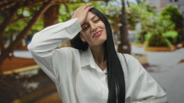 Güneşli bir parkta ağaçlar ve yeşilliklerle gülümseyen bir kadın, beyaz bir gömlek giymiş, rahat ve mutlu, huzurlu bir açık hava anını yakalıyor..