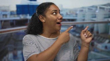 Gülümseyen kadın, bir binanın yanındaki gemi güvertesinde ufka doğru işaret ediyor; keyifli bir yolculuk.