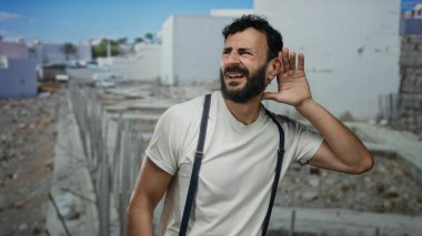 Sakallı İspanyol bir adam, açık mavi gökyüzünün altında bitmemiş binalarla çevrili bir inşaat alanında dikkatle dinliyor..