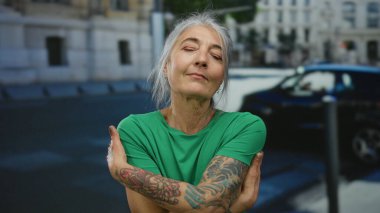 Dövmeli, yeşil tişörtlü, yaşlı bir kadın güneşli bir sokakta gözleri kapalı sarılıyor. Huzurlu bir yalnızlık anını yakalıyor..