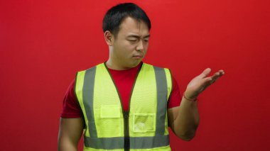 Yansıtıcı yelek giyen genç adam, kafa karışıklığı ve düşünceleri taşıyan canlı kırmızı bir zemin üzerinde duruyor..