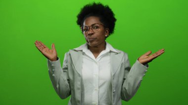 Gözlüklü ve gri ceketli, yeşil arka planda, elleri açık bir kadın omuz silkiyor..