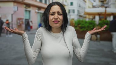 Beyaz gömlekli, uzun saçlı, kafası karışmış bir kadın. Renkli bir şehir zeminine karşı düşünceli görünüyor..