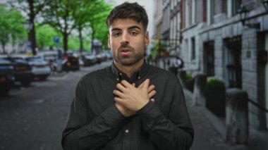 İspanyol bir adam göğsünü sıkıca tutarken iki eli kalabalık şehir caddesinde park halindeki arabaların yanında; korku endişesi.