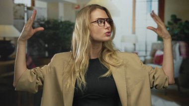 Zarif otel ortamında ellerini hareket ettiren kadın, gözlük ve bej ceket giyerken iletişim ve ifadeyi yansıtıyor..
