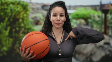 Parkta elinde basketbol topuyla gezen ve arka planda ağaçlarla kendini gösteren bir düdük takan genç İspanyol kadın.