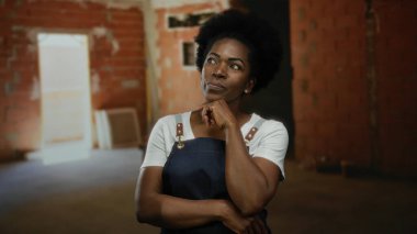 Önlüklü kadın düşünceli bir şekilde kapalı bir inşaat alanında duruyor, kollarını kavuşturmuş, planlama ve inşa etme konusunda güçlü ve düşünceli görünüyor..