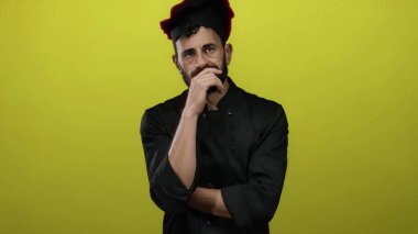 Siyah şef üniformalı İspanyol adam parlak sarı arka planda duruyor. Yorgun ve dalgın görünüyor. Yorgunluk ve yansıma duygularını aktarıyor..