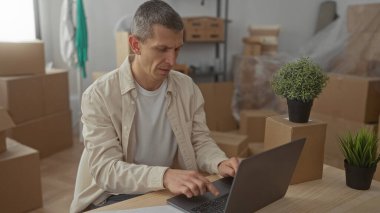 Dizüstü bilgisayarda çalışan sıradan giysili bir adam. Etrafı karton kutular ve bitkilerle çevrili. Yeni apartman dairesinde..