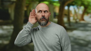 Sakallı ve kel kafalı bir adam, gri kazaklı, şehir caddesinde dikkatle dinliyor, bulanık bir şehir arka planına karşı tuhaf bir ifade sergiliyor..