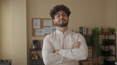 Sakallı genç İspanyol adam modern ofiste çapraz kollu, derin derin düşünüyor, kitaplarla ve belgelerle çevrili, tablolarla dolu bir tahtaya karşı..