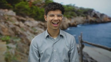 Gözlüklü bir adam açık mavi gökyüzü altında güneşli sahil şeridinde geniş bir gülümseme; tasasız mutluluk.