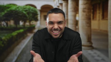 Erkek, taş sütunlar ve açık avuç içlerini gösteren yeşil çitlerle kaplı bir binanın avlusunda ellerini açar; merak bağlantısı açıklığı.