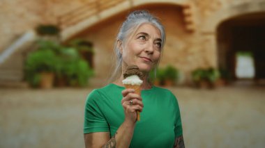 Eski üniversitede dondurmadan hoşlanan bir kadın, arka planda tarihi bir cazibeyle son sınıftaki bir bayanın huzurlu anını sergiliyor..