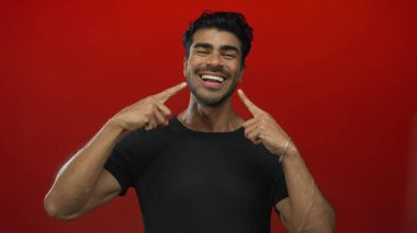 Siyah tişört giyen genç İspanyol adam kırmızı stüdyo ışıkları altında geniş bir gülümsemeyi işaret ediyor..