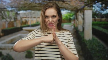 Avucunun içine sıkılmış bir yumruğu sıkıştıran kadın, güneşli bir parkta çitlerle ve taş bir çeşmeyle kaşlarını çatarken; öfke..