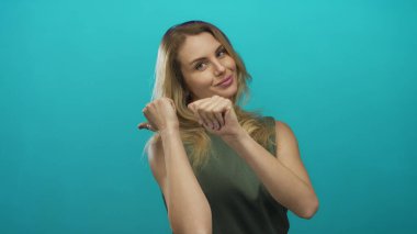 Başparmakları geriye doğru kıvrımlı yeşil arka planda gülümseyen kadın, izole edilmiş bir ortamda gençlik enerjisini, cazibesini ve pozitifliğini yakalar..