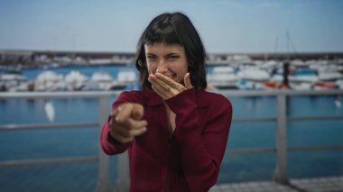Görünür dövmeli genç bir kadın ağzını kapatıyor ve demirli teknelerin yanındaki kamerayı işaret ediyor..