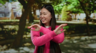 Canlı pembe bir kazakla gülümseyen kadın güzel yeşil bir parkta güneşli bir günün tadını çıkarıyor. Mutluluğunu doğa ve ağaçlarla ifade ediyor. Açık havada neşeli anlar sergiliyor..