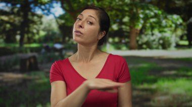 Orta yaşlı, kırmızı tişörtlü, yeşil parkta başını hafifçe eğmiş İspanyol bir kadın..