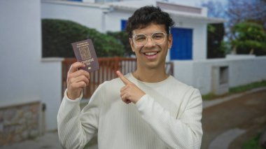 Beyaz binanın önündeki gülümseyen adam pasaport ve pasaportu tutuyor; seyahat heyecanı.