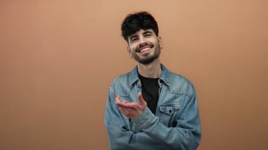Kot ceketli genç adam, düz kahverengi bir arka plan önünde kendinden emin bir şekilde gülümsüyor..