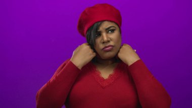 Kırmızı bereli ve gözleri kapalı bir kadın, canlı bir mor arka planda süveterini düzeltirken, ruh halini ve tarzını ön plana çıkarıyor..