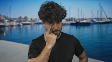 Güneşli bir günde arka planda tekne ve gemilerle dolu canlı bir limanda duran sakallı genç bir adam..