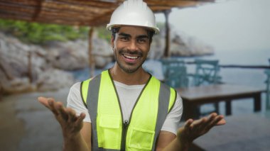 Neon yelek ve beyaz şapka giyen genç İspanyol adam deniz kenarı binasında el hareketi yapıyor; sıcak karşılama.