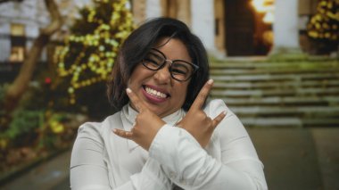 Gözlüklü genç İspanyol kadın şehir caddesinde rock jesti yapıyor. Arka planda neşeli ışıklar ve açık havada kendine güveni ifade ediyor..