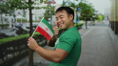 Başparmaklarıyla gülümseyen genç adam İran bayrağını şehir caddesinde tutuyor.