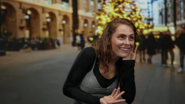 Genç bir kadın hareketli bir şehir sokağında kışın neşeli ışıklar ve bulanık kalabalıkla dinleyici jestler yapıyor..