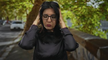 Siyah bluzlu kadın, yapraklı ağaçlarla dolu bir caddede elleri kulaklarına bastırıyor; stres gerilimi.
