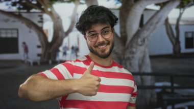 Sakallı ve gözlüklü genç adam arka planda ağaçlar ve binalar olan bir sokakta gülümsüyor kırmızı çizgili bir gömlek giyiyor..