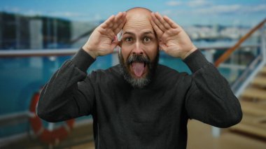 Sakallı, sakallı, komik bir şekilde yüzünü kapatan ve açık hava yolcu gemisinde mavi gökyüzü ve deniz altyapısı ile komik ifadeler kullanan, eşsiz bir ortamda dövmeleri olan kel bir adam..