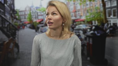 Genç sarışın kadın, park halindeki arabaların ve bina cephelerinin ortasında iki eliyle kulaklarını kapatıyor..