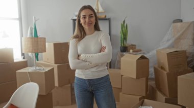 Karton kutularla çevrili yeni evinde duran genç bir kadın, kendine güvenle gülümsüyor.