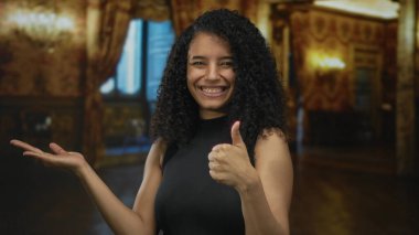 Latin kökenli genç bir kadın avucunun içini işaret ederek binanın içini işaret ederek mutluluğu onaylıyor..