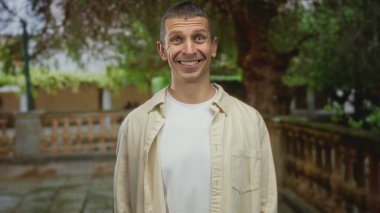 Ağaçlarla çevrili arka planda gülümseyen bir adam, tasasız, rahat, samimi bir an yakalıyor. Parlak ve renkli bir ortamda pozitif, rahat bir atmosfer tasvir ediyor..