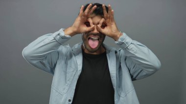 Kısa siyah saçlı İspanyol adam elleriyle dürbünlü jest yapıyor ve gri stüdyoda dilini gösteriyor..