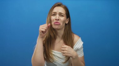 Beyaz omuzlu genç bir kadın dudaklarını büzüyor ve mavi stüdyo setinde yüzüne yumruk atıyor. Üzüntü..