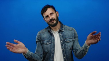 Sakallı, kot tişörtlü ve beyaz tişörtlü İspanyol bir adam mavi arka planda belirgin jestler yapıyor..