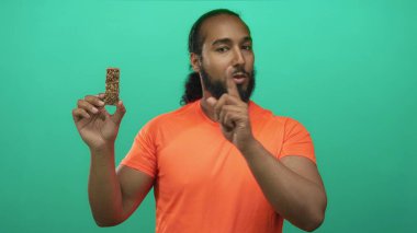 Parlak turuncu tişörtlü adam yeşil stüdyoda granola çubuğu tutuyor ve atıştırmak için parmağıyla işaret ediyor..