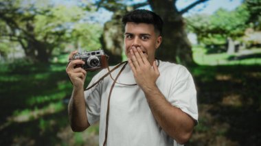 Parkta kameralı genç bir adam, yemyeşil ve parlak güneş ışığıyla çevrili şaşkınlığını ifade ediyor..