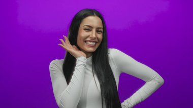 Genç İspanyol kadın neşeli bir dille gülümsüyor canlı mor bir arka planda, beyaz bir bluz giyiyor ve neşeli bir poz veriyor.