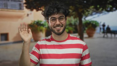 Sırıtan sakallı genç bir adam çizgili bir tişörtle sokakta ağaçlarla ve arka planda bir bankla durup sıradan ve dostane bir atmosfer yaratıyor.